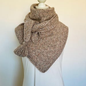 Può includere: Sciarpa lavorata a maglia marrone e beige, esposta su un manichino. La sciarpa presenta un collo alto e piegato e una forma triangolare, con un dettaglio a nappa. La trama della maglia è visibile, suggerendo un articolo fatto a mano e accogliente.