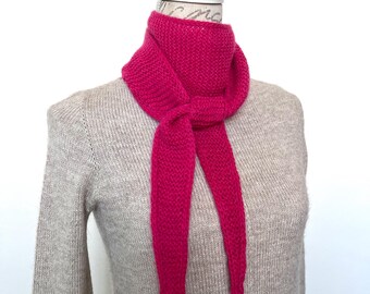May include: A bright pink knitted scarf with a long, narrow shape. The scarf is tied in a knot around the neck of a mannequin.