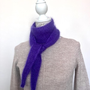 May include: A vibrant purple scarf is draped around a mannequin's neck. The scarf is made of a soft, fuzzy material and is knotted at the front. The mannequin is wearing a light brown sweater.