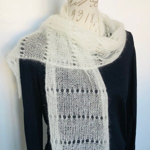 May include: A white, sheer, knitted scarf with a delicate, openwork lace pattern featuring horizontal rows of circular eyelets. The scarf has a soft, fuzzy texture and is draped around the neck of a mannequin wearing a solid black long-sleeve shirt.