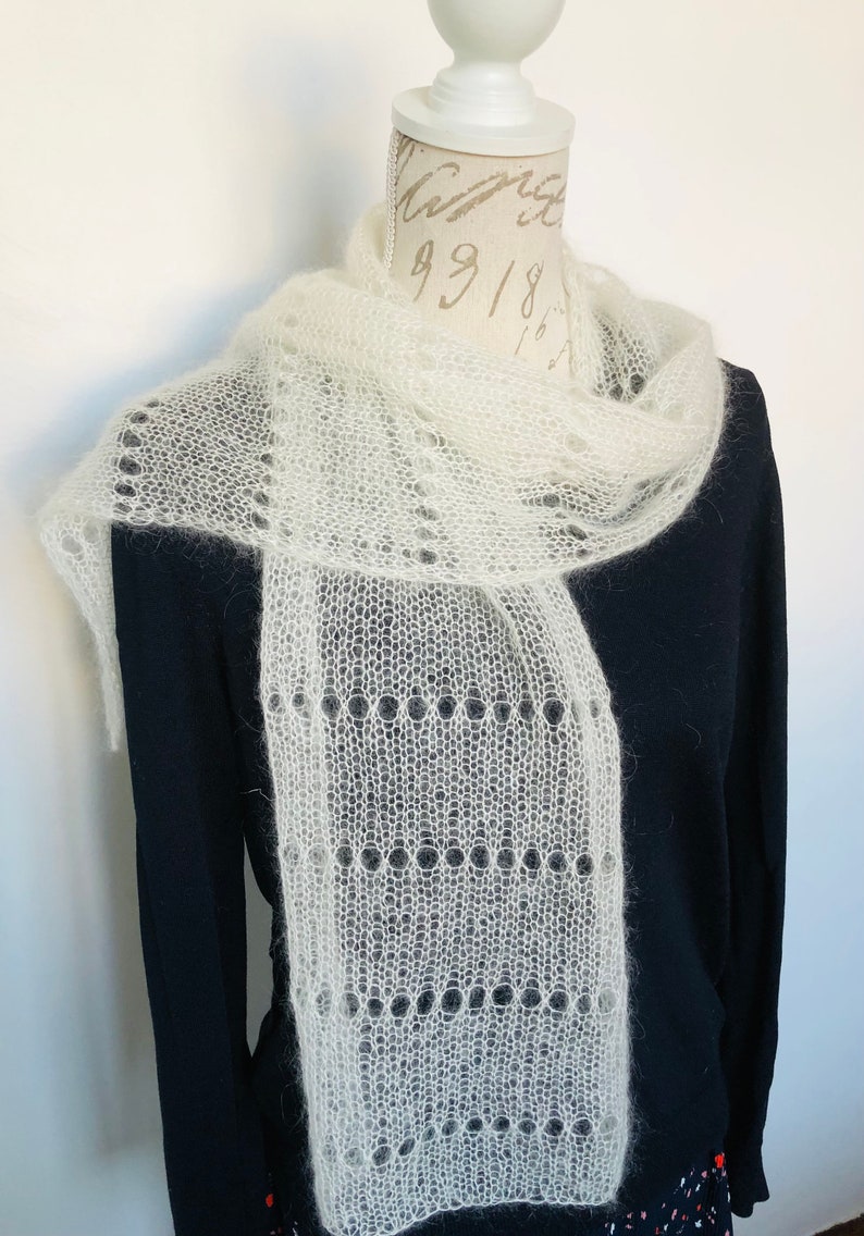 May include: A white, sheer, knitted scarf with a delicate, openwork lace pattern featuring horizontal rows of circular eyelets. The scarf has a soft, fuzzy texture and is draped around the neck of a mannequin wearing a solid black long-sleeve shirt.