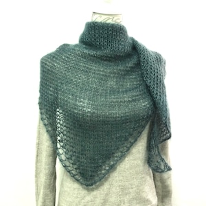 Pode incluir: Um xale triangular verde-azulado escuro com uma textura de malha solta e aberta, apresentando uma aparência felpuda semelhante ao mohair. O xale está drapeado sobre um suéter cinza-claro de manga comprida, exibindo uma forma larga e pontiaguda.