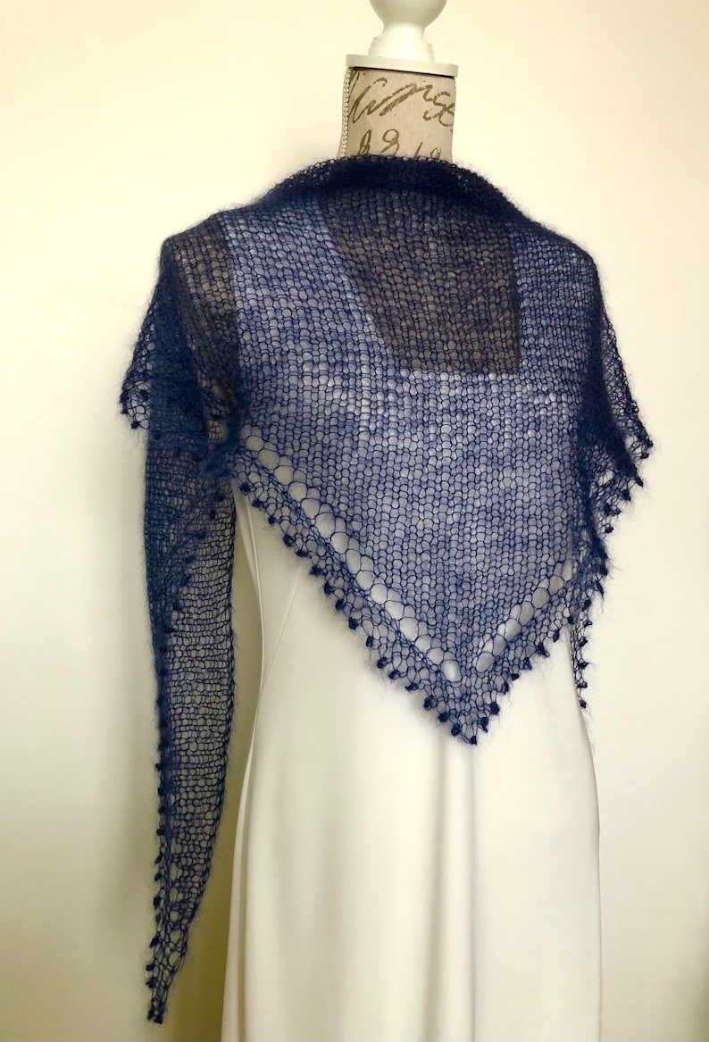 May include: A dark navy blue triangular shawl featuring a loose, open-knit texture with a fuzzy appearance. The edges of the shawl are finished with a decorative border of small, dangling loops, creating a delicate and airy look for this accessory.