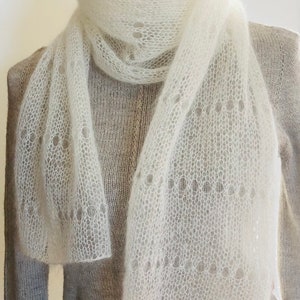 May include: A white, semi-sheer, knitted scarf with a delicate, open-work lace pattern featuring circular motifs. The scarf is draped around the neck of a mannequin wearing a light beige, long-sleeved, knit sweater with a visible vertical seam.