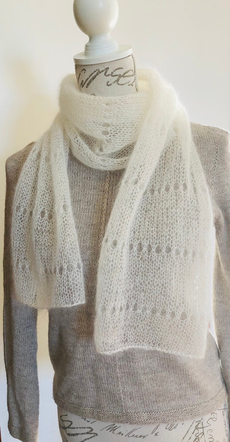 May include: A white, semi-sheer, knitted scarf with a delicate, open-work lace pattern featuring circular motifs. The scarf is draped around the neck of a mannequin wearing a light beige, long-sleeved, knit sweater with a visible vertical seam.