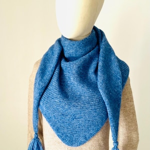 Puede incluir: Una bufanda triangular tejida en un vibrante color azul medio. La bufanda presenta un patrón de punto bobo texturizado en toda su superficie y está rematada con dos borlas de hilo azul a juego unidas a las esquinas inferiores de la forma.