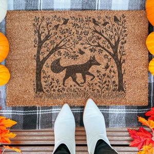 Op de afbeelding: Een bruine kokosvezel deurmat met een zwart silhouet van een vos die door een bos loopt met vogels en bomen. De deurmat ligt op een zwart-wit geblokt oppervlak.