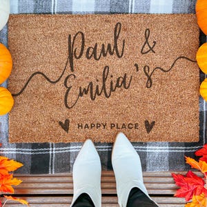 May include: A brown coir doormat with the text "Paul & Emilia's Happy Place" in black lettering. The doormat is on a black and white checked surface.