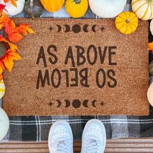May include: A rectangular coir doormat with the text "AS ABOVE SO BELOW" in black. The mat is surrounded by pumpkins, gourds, and autumn leaves. White trainers are visible at the bottom of the image.