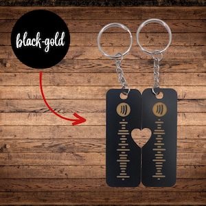 Op de afbeelding: Twee zwarte sleutelhangers met goudkleurige Spotify-code ontwerpen. Eén sleutelhanger heeft een hartvormige uitsparing waar de twee sleutelhangers samenkomen. De tekst "black-gold" staat in een zwarte cirkel linksboven.