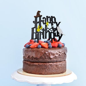 May include: A two-tier chocolate cake decorated with fresh strawberries, blueberries, and a black "Happy Birthday" cake topper with a wizard theme. The cake is on a white cake stand against a light blue backdrop.
