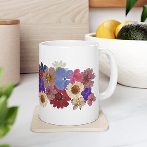 Puede incluir: Taza de cerámica blanca decorada con un colorido arreglo floral de flores prensadas. Las flores incluyen tonos de rojo, morado, azul y amarillo. La taza está sobre un posavasos beige.