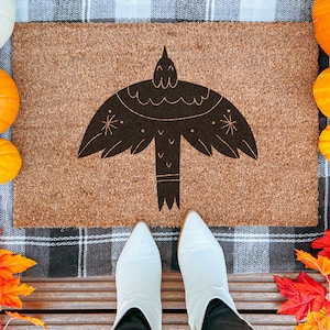 May include: A rectangular doormat with a brown background and a black bird design. The bird has outstretched wings and decorative details. The mat is surrounded by pumpkins and fall leaves, with white boots in the foreground.