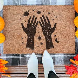 Peut inclure: Un paillasson rectangulaire en fibre de coco naturelle avec un motif céleste. Deux mains tendues noires sont entourées d'étoiles, d'une planète et d'un croissant de lune. Des citrouilles et des feuilles d'automne entourent le tapis.