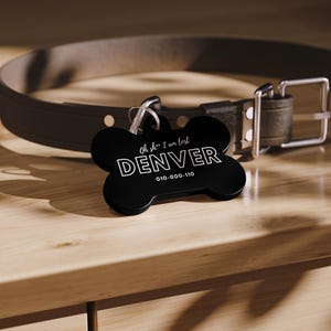 Op de afbeelding: Zwarte, botvormige hondenpenning met witte tekst die "Oh sh*... I am lost DENVER 010-000-110" luidt.