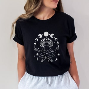 Op de afbeelding: Zwart T-shirt met een witte afbeelding van een mediterende kikker met een paddenstoelkap, omringd door halve manen en sterren. De kikker zit in de lotushouding met uitgestrekte armen. Het shirt heeft korte mouwen.