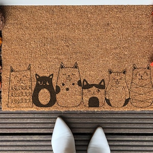 Könnte beinhalten: Eine braune Kokosfaser-Fußmatte mit einer Reihe von sechs schwarzen und weißen Katzen mit verschiedenen Gesichtsausdrücken. Die Katzen sind in einem einfachen, cartoonartigen Stil gezeichnet. Die Fußmatte liegt auf einer Holzoberfläche.