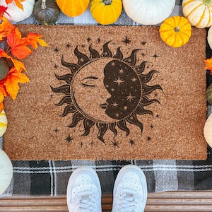 Op de afbeelding: Een bruine kokosdeurmat met een hemels ontwerp van een zon en maan met sterren. De zon en maan zijn donkerbruin. De deurmat is omgeven door pompoenen en herfstbladeren.