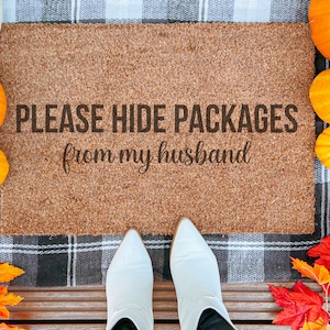 May include: A rectangular coir doormat with the text "PLEASE HIDE PACKAGES from my husband" in black lettering. The mat is surrounded by pumpkins, gourds, and fall foliage. White boots are visible at the bottom of the image.