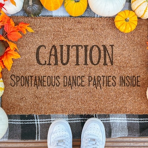 May include: A rectangular doormat with the text "CAUTION SPONTANEOUS DANCE PARTIES INSIDE". The mat is surrounded by pumpkins in various colors and fall foliage. White sneakers are at the bottom.
