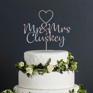 Puede incluir: Una tarta de boda blanca de tres pisos con una guirnalda floral y un adorno que dice "Mrs & Mrs Cluskey" con un corazón encima. La tarta tiene un patrón acolchado en el nivel medio.