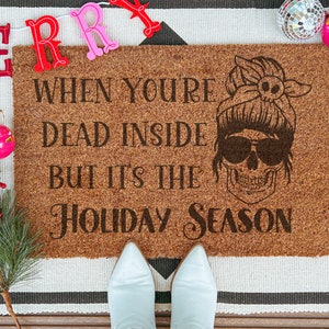 Può includere: Un zerbino in fibra di cocco marrone con la scritta "WHEN YOU'RE DEAD INSIDE BUT IT'S THE HOLIDAY SEASON" e una grafica a teschio con occhiali da sole. Decorazioni festive con uno striscione "MERRY", ornamenti e una ghirlanda.