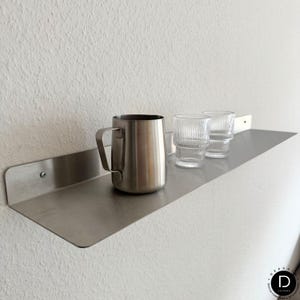 May include: A brushed stainless steel wall-mounted shelf featuring a single metal mug and two clear, vertically ribbed glass tumblers. The shelf has a flat horizontal surface and a vertical back panel with visible screw holes for mounting.