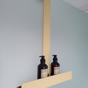 May include: A gold-colored metal over-the-door shower caddy featuring a long vertical support arm and a horizontal shelf. Two dark brown plastic pump bottles with labels reading meraki are placed on the shelf, containing body wash and body lotion.
