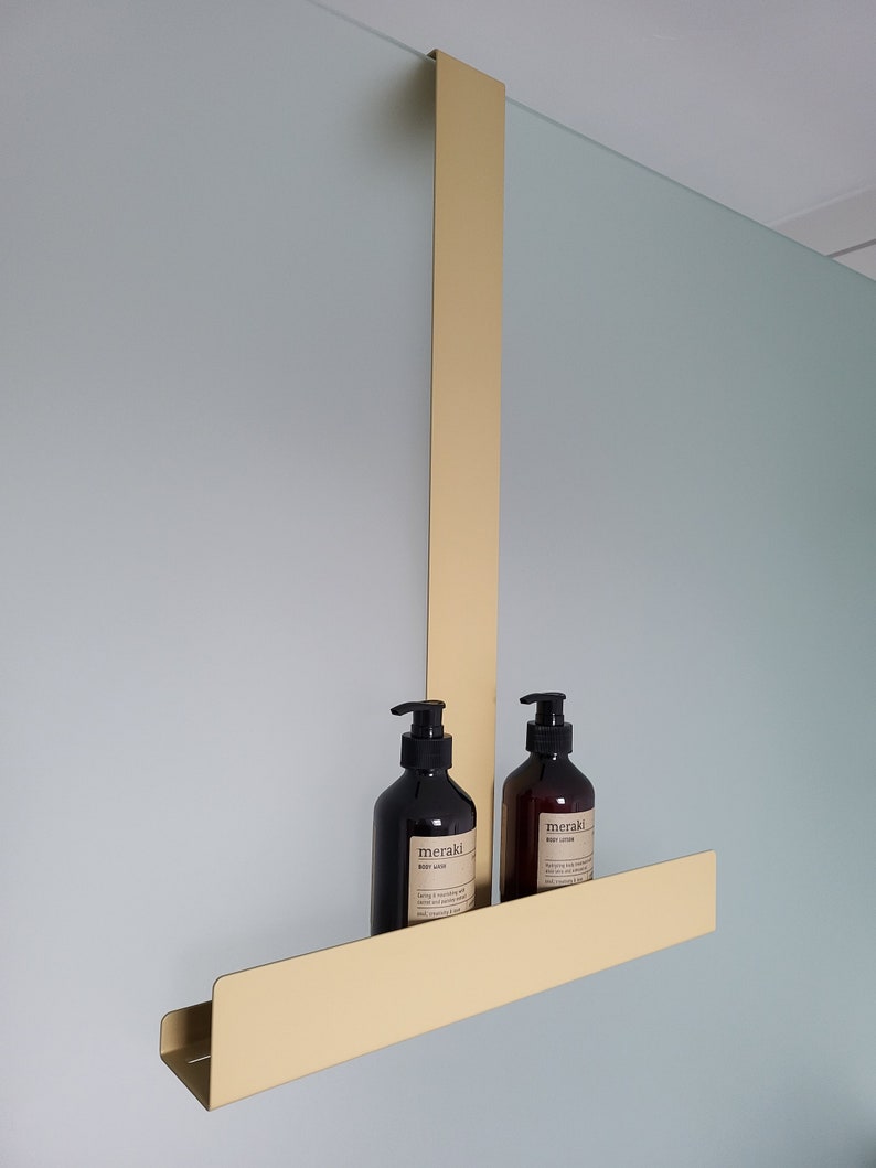 May include: A gold-colored metal over-the-door shower caddy featuring a long vertical support arm and a horizontal shelf. Two dark brown plastic pump bottles with labels reading meraki are placed on the shelf, containing body wash and body lotion.