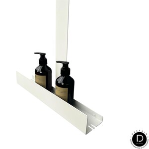 May include: A white metal wall-mounted shelf with a vertical back panel, holding two dark brown plastic pump bottles with beige labels. The shelf features a long, narrow tray design with a small cutout detail on the right side of the base.