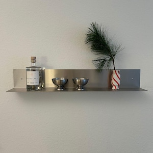 May include: A brushed metal wall shelf holding a clear glass bottle with a Woodland label, two small stainless steel pedestal bowls, and a white vase with red diagonal stripes containing a green pine branch with long, thin needles.