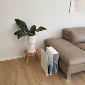 Könnte beinhalten: Weißer Magazinhalter aus Metall mit zwei Fächern, in denen Bücher und Zeitschriften liegen. Der Halter steht auf einem Holzhocker neben einem hellbraunen Sofa. Ein weißer Keramiktopf mit einer grünen Pflanze steht auf dem Hocker.
