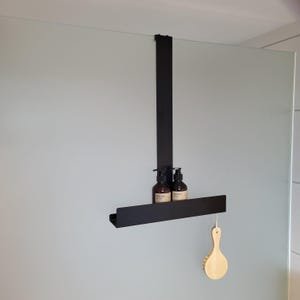 May include: A matte black metal over-the-glass shower caddy featuring a single horizontal shelf. Two amber-colored pump bottles with beige labels are placed on the shelf, and a light-colored wooden bath brush hangs from the right side of the frame.