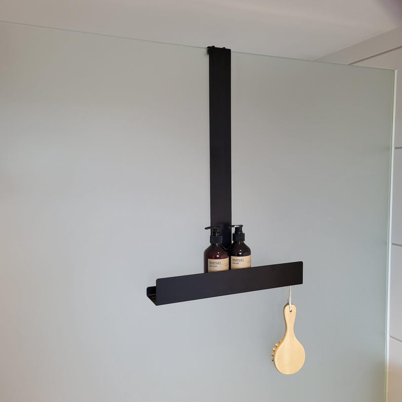 May include: A matte black metal over-the-glass shower caddy featuring a single horizontal shelf. Two amber-colored pump bottles with beige labels are placed on the shelf, and a light-colored wooden bath brush hangs from the right side of the frame.
