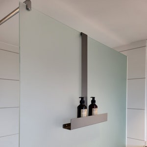 May include: A matte dark gray metal shower caddy hanging over a frosted glass partition. The caddy features a long vertical support arm and a horizontal shelf holding two dark brown plastic pump bottles with white labels and black pump tops.