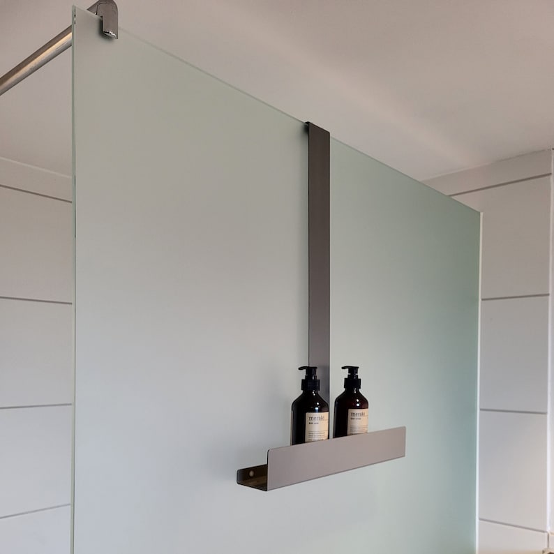May include: A matte dark gray metal shower caddy hanging over a frosted glass partition. The caddy features a long vertical support arm and a horizontal shelf holding two dark brown plastic pump bottles with white labels and black pump tops.