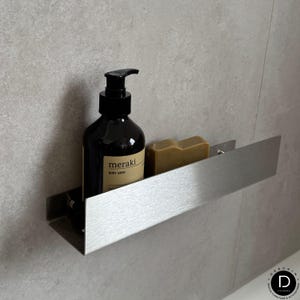 May include: A stainless steel bathroom shelf holding a dark brown bottle of "meraki BODY WASH" and a bar of soap. The shelf is mounted on a gray tiled wall. The shelf is approximately 12 inches long.