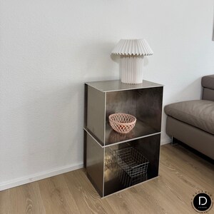 Könnte beinhalten: Ein moderner, zweistöckiger Beistelltisch aus Metall mit einer weißen, plissierten Lampe. Auf dem oberen Regal befindet sich ein kleiner, rosa gewebter Korb und auf dem unteren Regal ein Drahtkorb. Der Tisch steht in einem Raum mit neutraler Farbgebung.