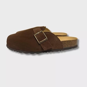 Op de afbeelding: Bruine suède klompen met een kurken voetbed en een gespsluiting. De klompen hebben een gesloten neusontwerp en een donkerbruine rubberen zool. De gesp is vierkant en gemaakt van goudkleurig metaal.