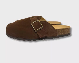 May include: Brown suede clogs with a cork footbed and a buckle closure. The clogs have a closed-toe design and a dark brown rubber sole. The buckle is a square shape and is made of a gold-colored metal.
