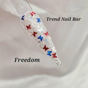 May include: A white nail with red, white, and blue butterfly nail art. The nail art is on a white background with the text "Trend Nail Bar" and "Freedom".