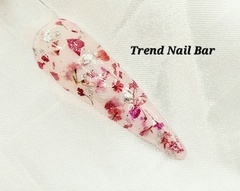 Puede incluir: Primer plano de una uña larga y transparente con base rosa y un diseño de flores secas y purpurina plateada. El texto "Trend Nail Bar" es visible en el fondo.