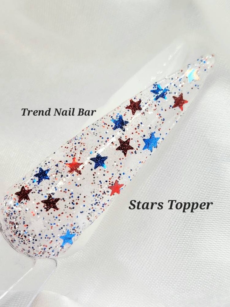 May include: A clear nail polish topper with red and blue glitter stars. The text "Trend Nail Bar" and "Stars Topper" are visible in the image.