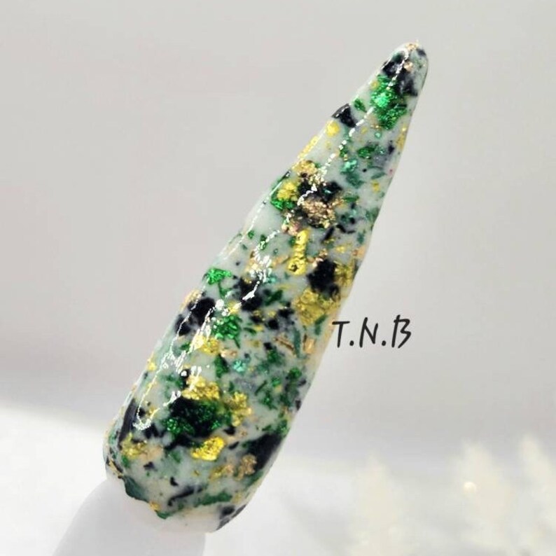 Pu&ograve; includere: Uno smalto per unghie bianco con glitter verde, oro e nero. Lo smalto per unghie &egrave; su una punta di unghia bianca. Il testo "T.N.B" &egrave; scritto sull'immagine.
