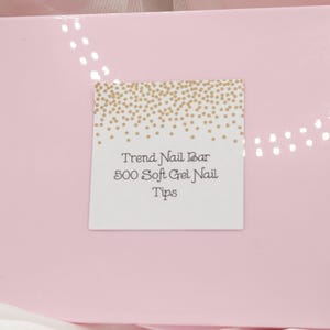 May include: A pink box with a white label featuring the text "Trend Nail Bar 500 Soft Gel Nail Tips." The label has a gold confetti design at the top. The box is a light pink color.