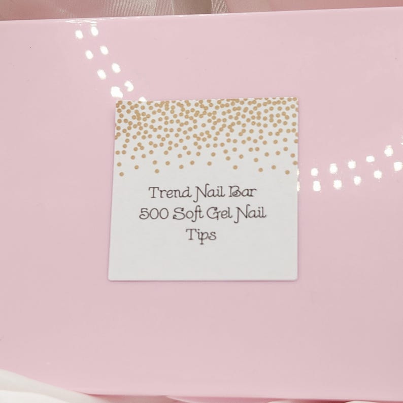 May include: A pink box with a white label featuring the text "Trend Nail Bar 500 Soft Gel Nail Tips." The label has a gold confetti design at the top. The box is a light pink color.