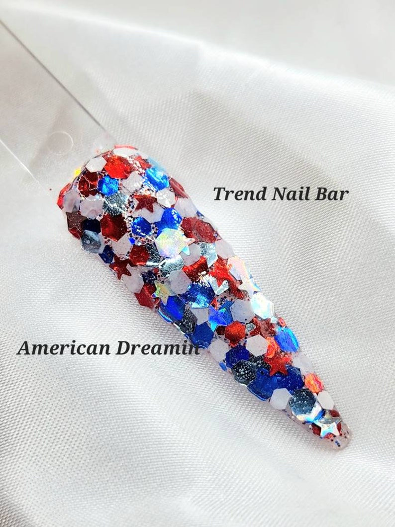 May include: Red, white, and blue glitter nail polish with a star-shaped design. The polish is called "American Dreamin'" and is from Trend Nail Bar.