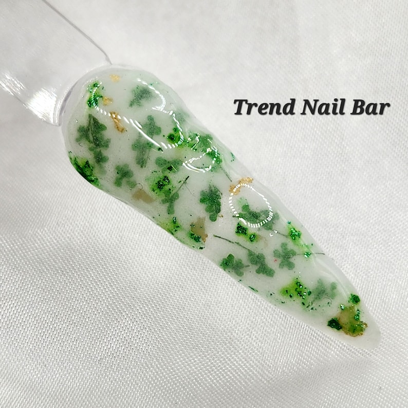 K&ouml;nnte beinhalten: Nahaufnahme eines transparenten Nagelspitzen mit gr&uuml;nem und goldenem Glitzer, eingebettet in den klaren Lack. Der Text "Trend Nail Bar" ist in der oberen rechten Ecke des Bildes sichtbar.