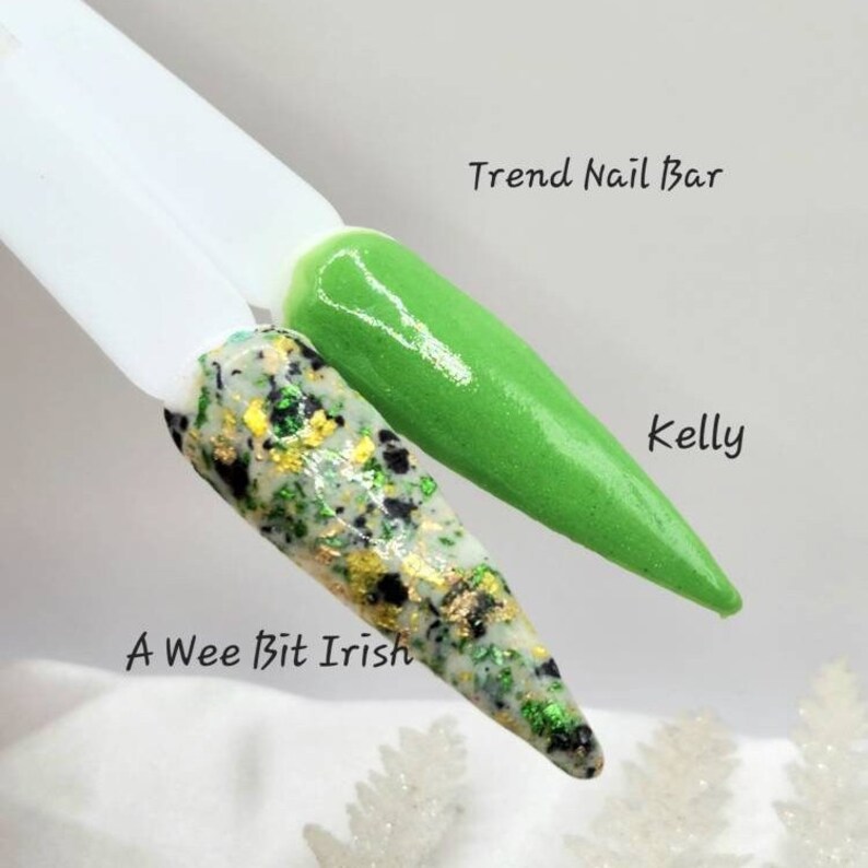 Pu&ograve; includere: Due flaconi di smalto per unghie, uno verde e uno con una finitura glitter verde, nero e oro. Il flacone verde &egrave; etichettato "Kelly" e il flacone glitter &egrave; etichettato "A Wee Bit Irish". I flaconi sono su una superficie bianca con il testo "Trend Nail Bar" sopra.