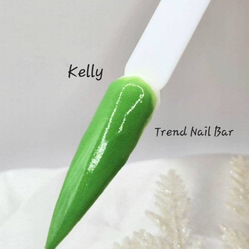 Pu&ograve; includere: Primo piano di uno smalto per unghie verde su una spatola bianca. Il testo "Kelly" &egrave; scritto sopra la spatola e "Trend Nail Bar" &egrave; scritto sotto la spatola.